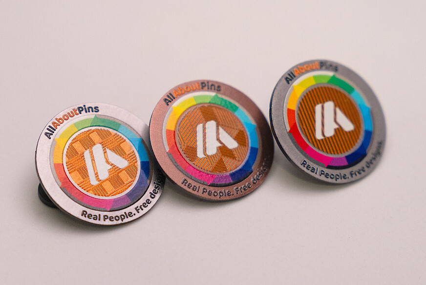 Three round printed pins in white, copper and silver metal with rainbow rings and orange centers, on a light gray surface