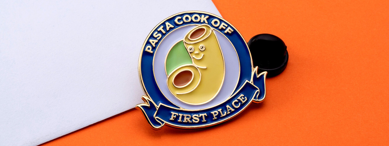 Blue and gold Pasta Cook Off First Place pin with a smiling noodle, beside a black clutch on orange paper