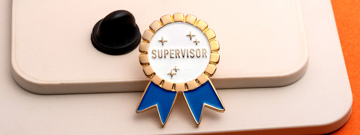 White and gold rosette pin reading Supervisor with blue ribbon tails, beside a black clutch