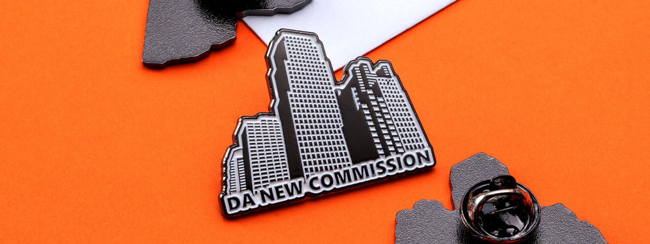 Black and silver skyscraper pin reading DA New Commission, beside its clutch on orange paper