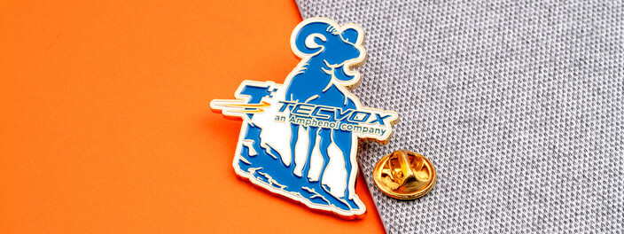 Blue ram mascot pin with a gold Tecvox name bar, beside a gold clutch on gray fabric