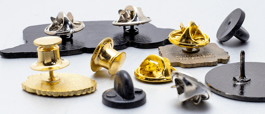 Gold, silver and black butterfly clutches, locking backs and rubber clutches scattered around pin backs on a white surface
