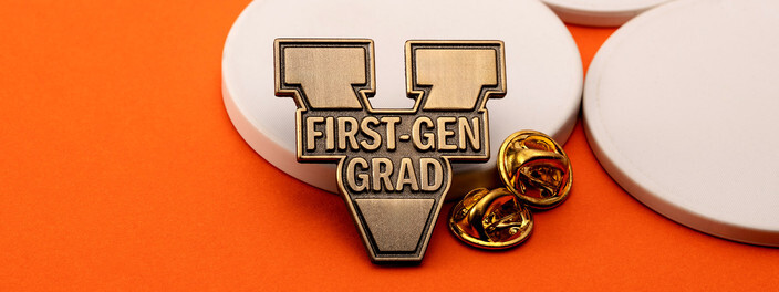 Antique brass V pin reading First-Gen Grad, beside two gold clutches on white discs