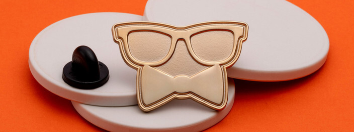 Gold pin shaped like a pair of glasses over a bow tie, on white discs beside a black clutch