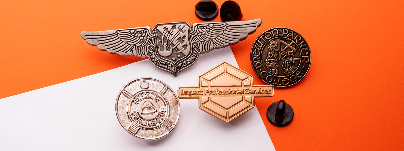 Silver winged pin above round and hexagon metal pins in gold, silver and copper, with their clutches on orange paper