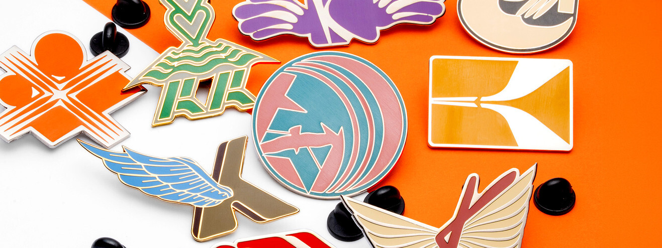 Pile of bright hard enamel pins in many shapes, among them a globe, a pair of wings and a pink logo, on orange paper