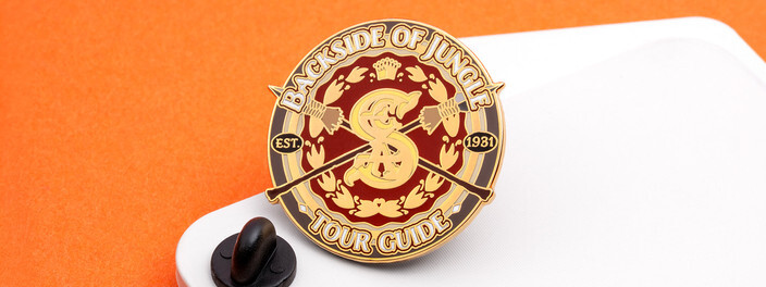 Round gold tour guide pin with a jungle explorer emblem, beside a black clutch on a white board