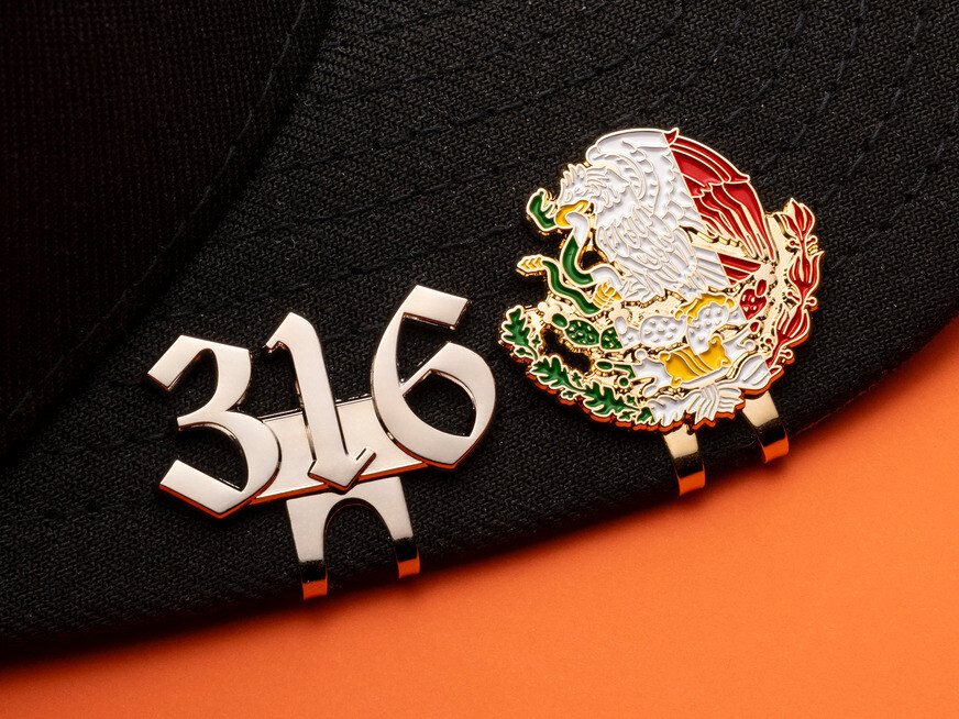 Silver 316 number clip and a gold eagle crest clip in red, white and green enamel on a black cap brim over orange paper