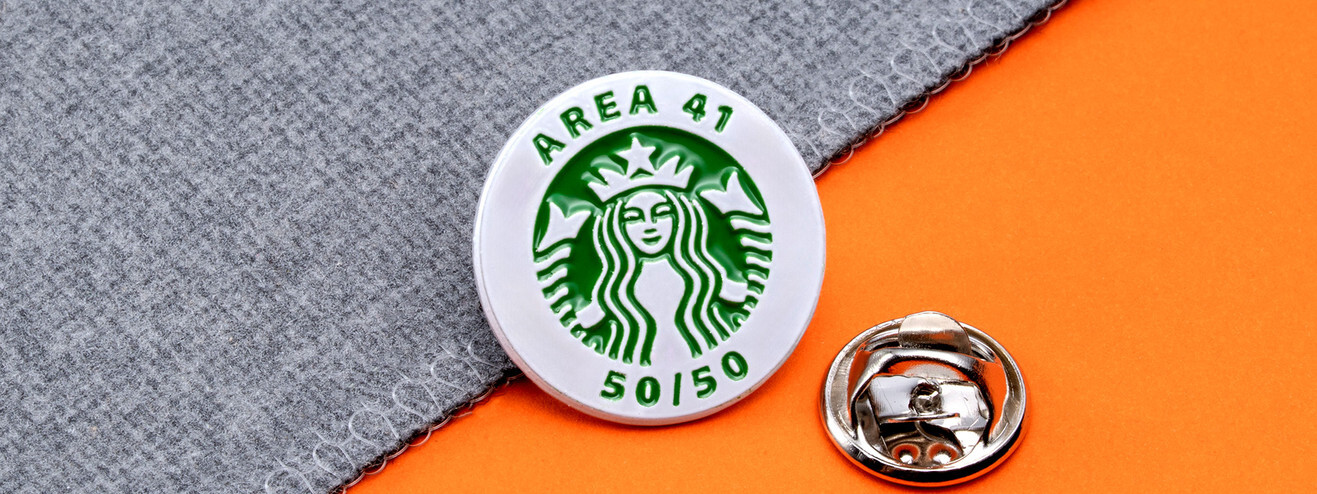 Round white pin with a green siren logo, Area 41 and 50/50, beside a silver clutch on gray fabric and orange paper