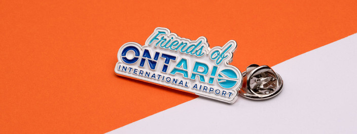 Silver pin reading Friends of Ontario International Airport in blue and teal, beside its clutch on orange and white paper