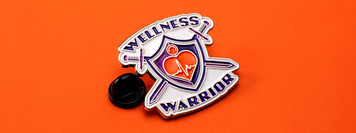 Wellness Warrior pin with a purple shield and a red heart, beside a black clutch on orange paper