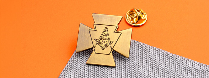 Antique bronze cross shaped pin with a keystone holding the square and compasses, beside a gold clutch on orange paper