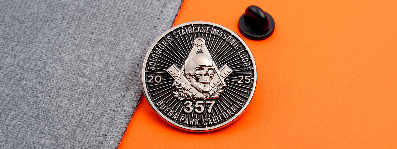 Antique silver Masonic lodge pin with a skull over the square and compasses, dated 2025, beside a black clutch on gray fabric