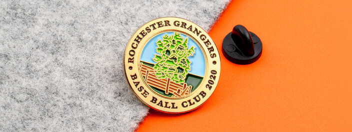 Gold rimmed base ball club 2020 pin with a green tree behind a wooden fence, beside a black clutch