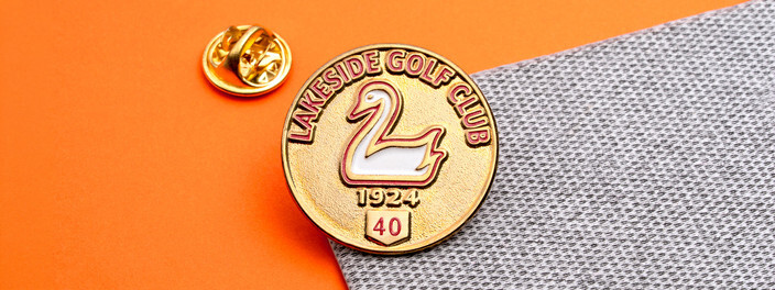Gold round golf club pin with a white swan, the year 1924 and a red 40 badge, beside a gold clutch