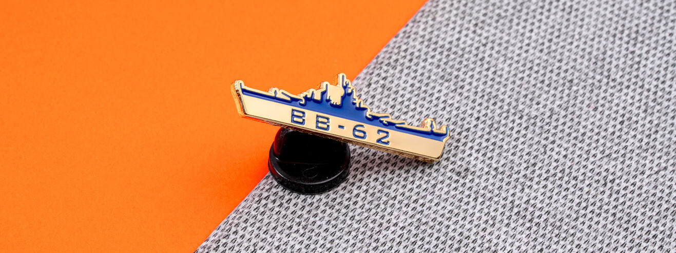 Blue and gold battleship silhouette pin reading BB-62 on its black clutch, on orange paper and gray fabric