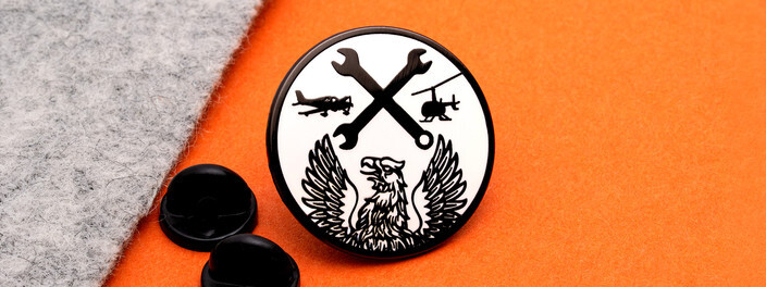 Round black and white pin with crossed wrenches between an airplane and a helicopter over a phoenix, beside two black clutches
