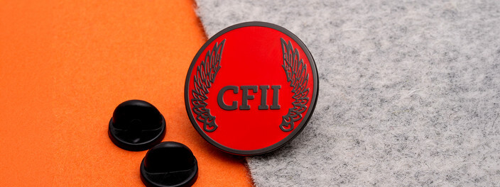 Round red pin reading CFII between two black wings, beside two black clutches on orange paper and gray felt