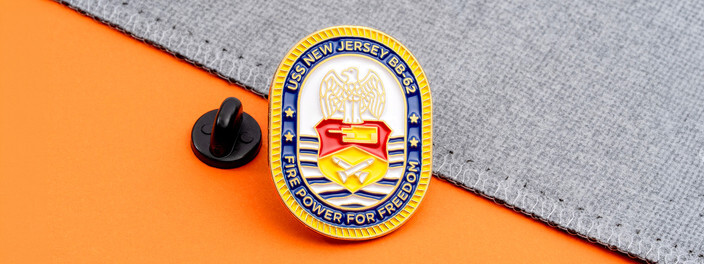 Gold rimmed USS New Jersey BB-62 crest pin with an eagle and Fire Power for Freedom, beside a black clutch on orange paper