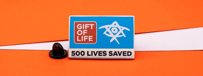 Rectangular blue pin reading 500 Lives Saved, beside a black clutch on orange and white paper