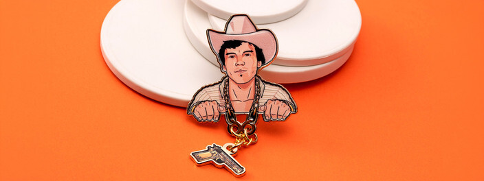 Caricature pin of a man in a pink cowboy hat gripping a gold chain, with a small pistol charm hanging below, on white discs and orange paper