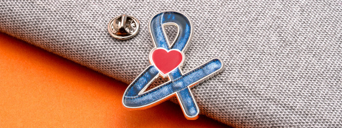 Awareness ribbon pin printed with blue denim and a red heart where the loops cross, beside its clutch on gray fabric and orange paper