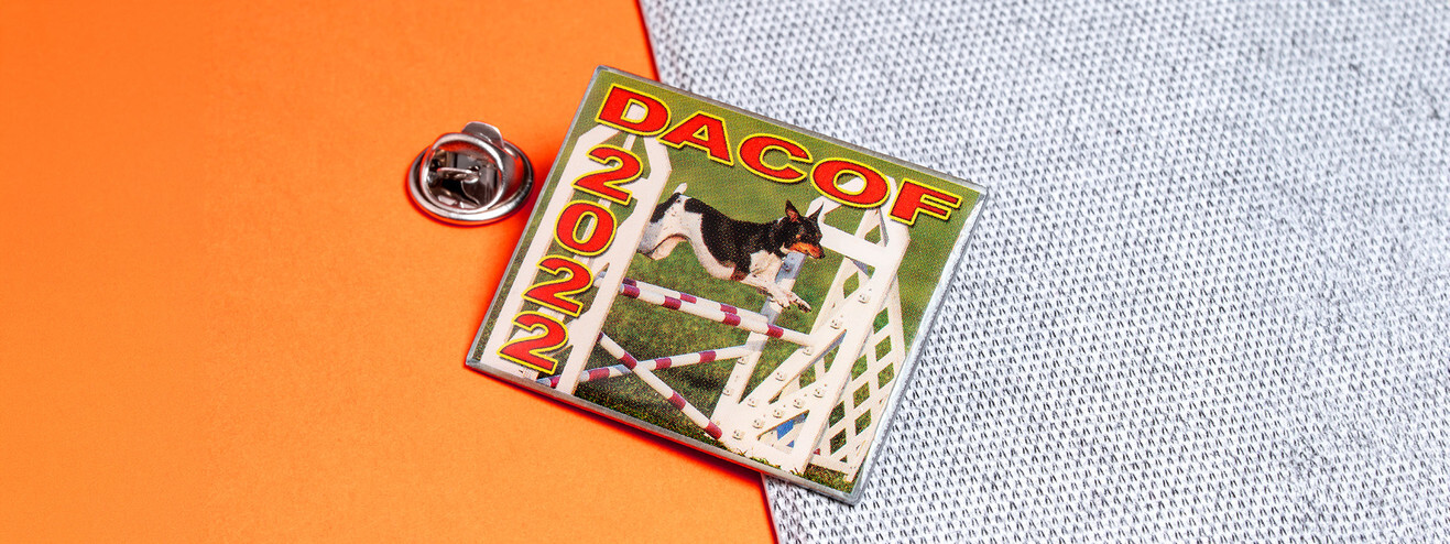 Square pin with a photo of a small dog leaping an agility jump under DACOF 2022 in red letters, beside its clutch on orange paper and gray fabric