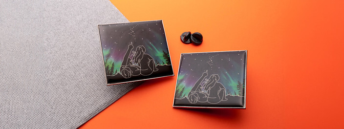 Two square pins of white-outlined figures under green northern lights and stars, beside two black clutches on gray fabric and orange paper
