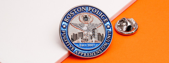 Round blue and silver fugitive apprehension unit pin with an eagle over a city skyline, beside a silver clutch on orange paper