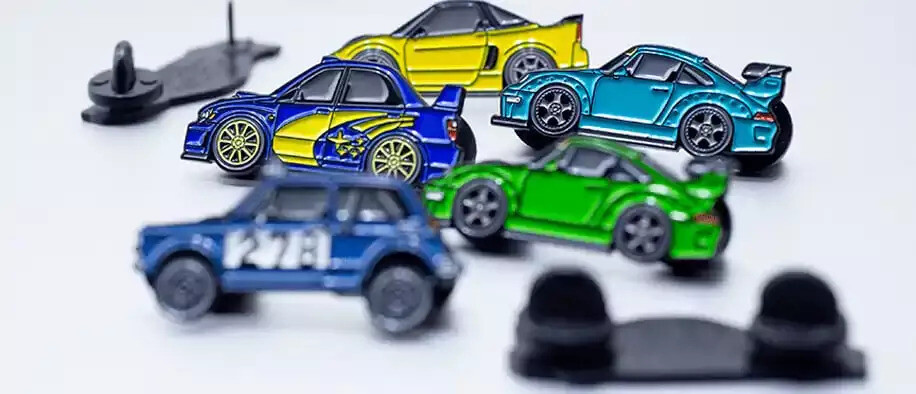 Small enamel car pins in yellow, blue, teal and green standing on a white surface beside black rubber clutches