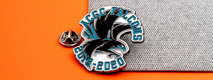 Black and teal falcon pin reading Falcons 2019-2020, beside a silver clutch on gray fabric