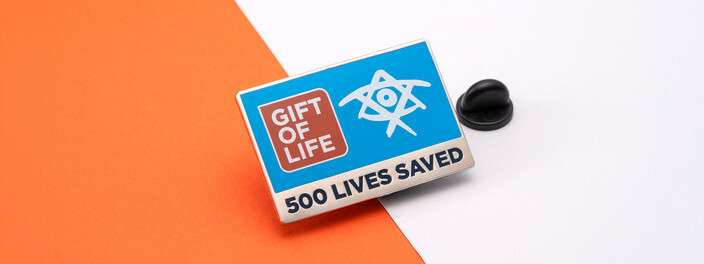 Blue rectangular Gift of Life pin reading 500 Lives Saved, beside a black clutch on orange and white paper