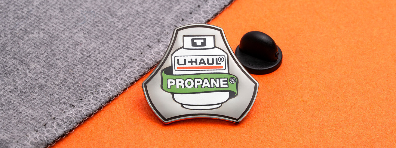 Pin shaped like a propane tank with a U-Haul label and a green Propane banner, beside a black clutch on orange paper and gray fabric