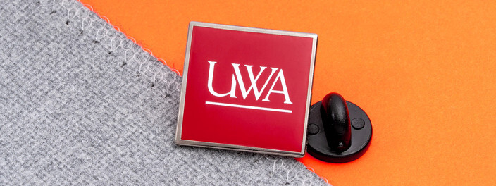Square crimson pin with a white UWA wordmark, beside a black clutch on gray fabric and orange paper