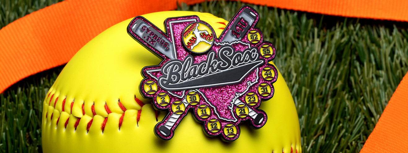 Pink glitter Black Sox softball trading pin with crossed bats and a ring of numbered softballs, resting on a softball on turf